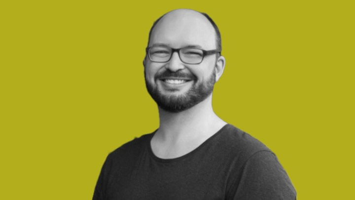 Chris Simon in black & white, smiling at the camera in front of a grokludo yellow background.